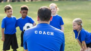 In an outside setting, a sports coach has his back to the camera and we can see the word COACH across his top as four children gather round with one holding a football to represent this to represent this blog about the best children's sports franchise businesses for sale in the UK