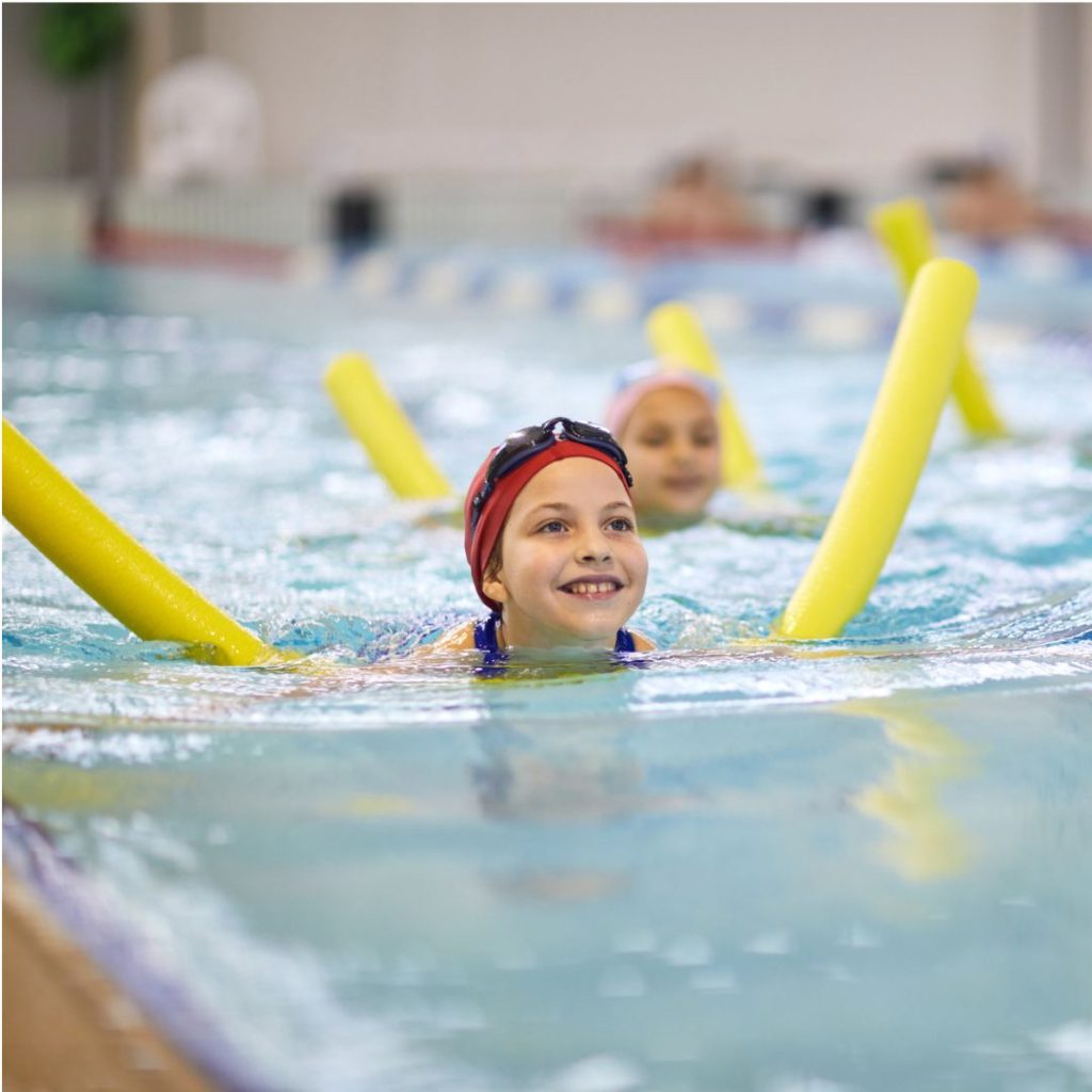 A young girl smiles as she enjoys a swimming lesson in a large indoor pool to represent this blog about the best children's sports franchise businesses for sale in the UK