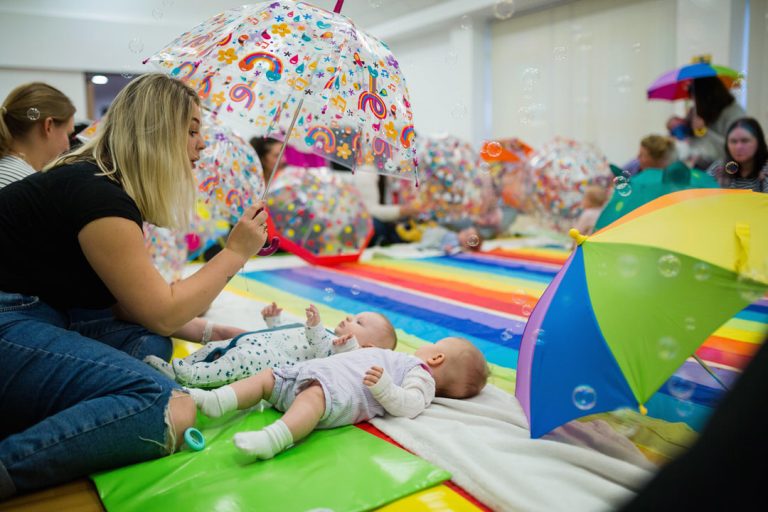 The Baby Sensory class in action with a group of babies laying on foam floor mats. Indoor umbrellas and colourful props are held above them to aid development and entertain them.