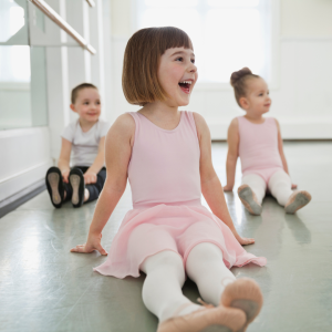 Three young girls in ballet outfits sit on the floor of a dance studio with big smiles to represent this blog about the best children's dance franchise businesses for sale in the UK
