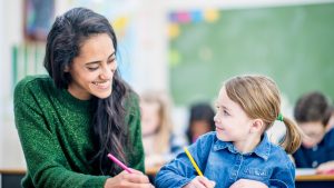 A teacher sits at a desk next to a young girl in a classroom and helps her with her studies to represent this blog about franchise career opportunities for teachers thinking about leaving teaching