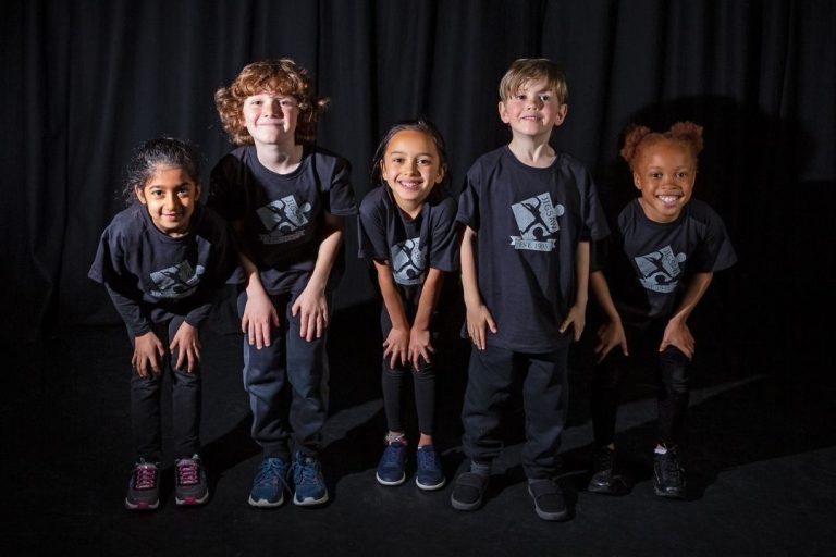 Children from Jigsaw Performing Arts smile and pose in their branded t-shirts to promote this successful drama franchise