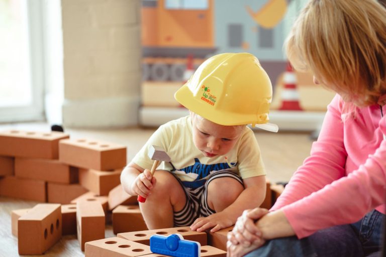 A young boy plays amongst a toy building site wearing a yellow hardhat as his mother watches on at a role play session from the Pop Up Play Village franchise