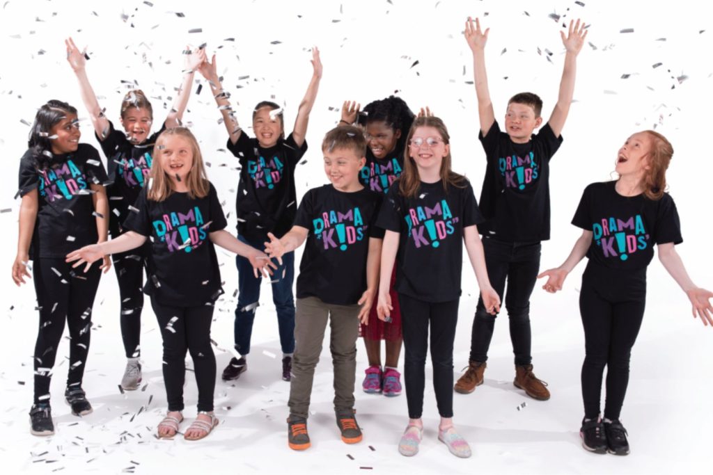 A group of children raise their arms in the air surrounded by falling confetti and wearing branded t-shirts in celebration of the Drama Kids global drama franchise brand