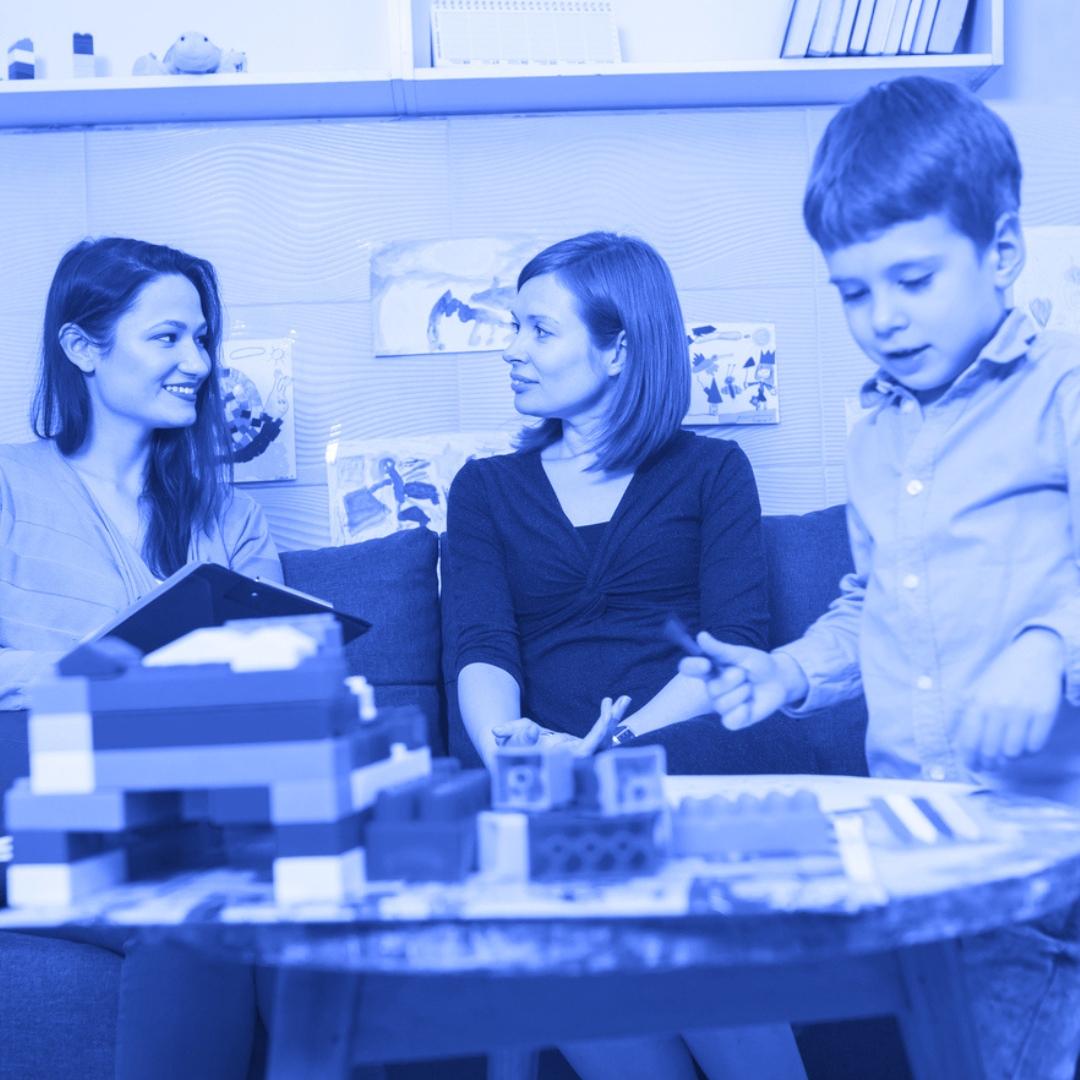 An educator and parent sit together while a young boy plays at a table with plastic bricks and felt pens in an image that represents the National College for Care and Education partnership opportunity