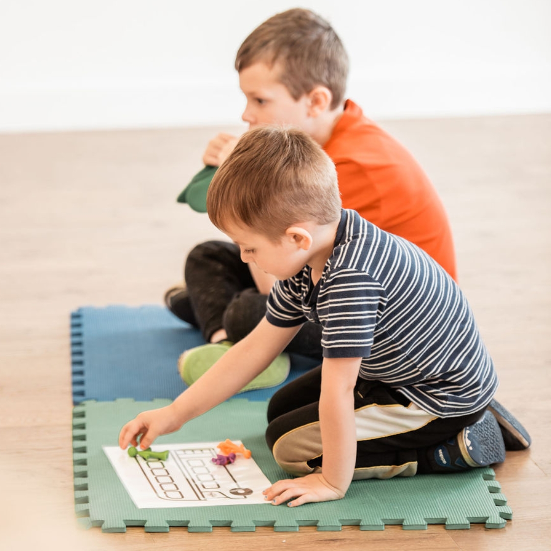 Two young boys sit together on a mat and work with colourful plastic figures and a drawing of a bus at a fun maths session with the franchise team at Counting Kids