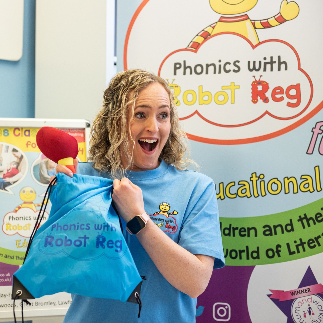 A smiling team member from the Phonics with Robot Reg franchise holds up a branded bag and is about to pull out a Reg robot soft toy at a fun phonics sessions