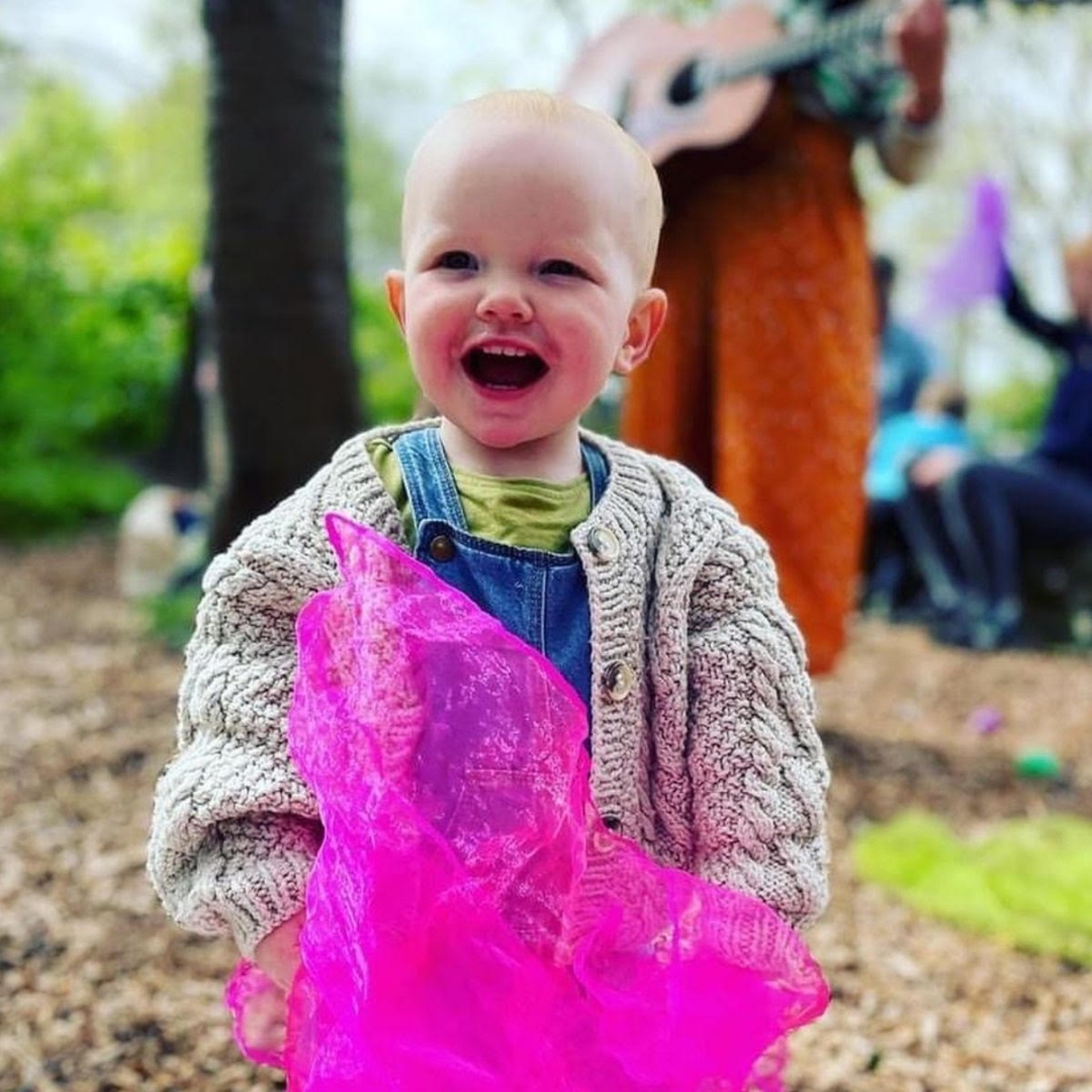 A young child happily enjoys an outdoor session waving a colourful scarf prop as a leader plays guitar at music franchise Rock Out Baby