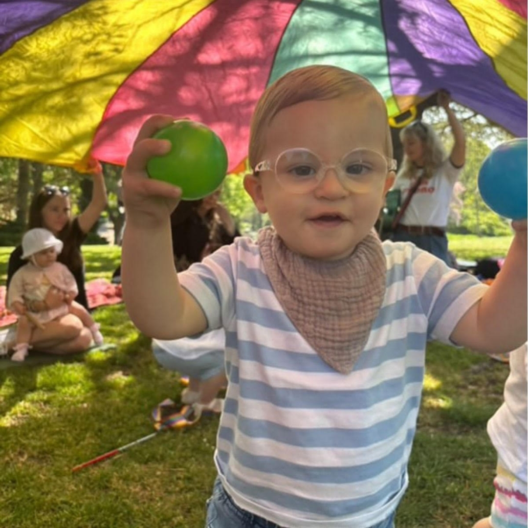 A young boy enjoys playing with sensory balls beneath a colourful parachute in a sunny outdoor setting at a session with children's music franchise Rock Out Baby