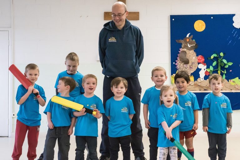A group of young children stand and smile while holding cricket bats and wearing branded t-shirts in front of their coach at a fun session with children's sports franchise brand Little Wickets