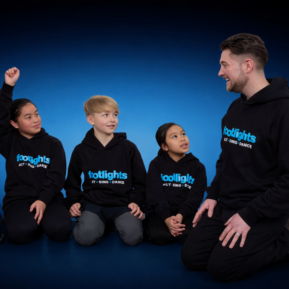 Three young children sit on the floor and engage enthusiastically with a tutor from Footlights the children's drama franchise UK