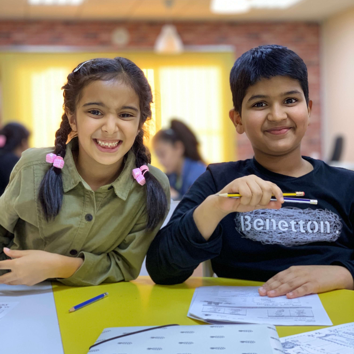 A young boy and a young girl smile happily as they look up from their worksheets at a tuition session with MagiKats, the children's education franchise UK